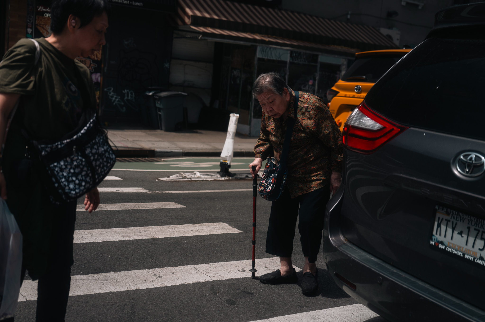 A street crossing near the shop in harsh morning sun, an elderly woman with a cane passing through.