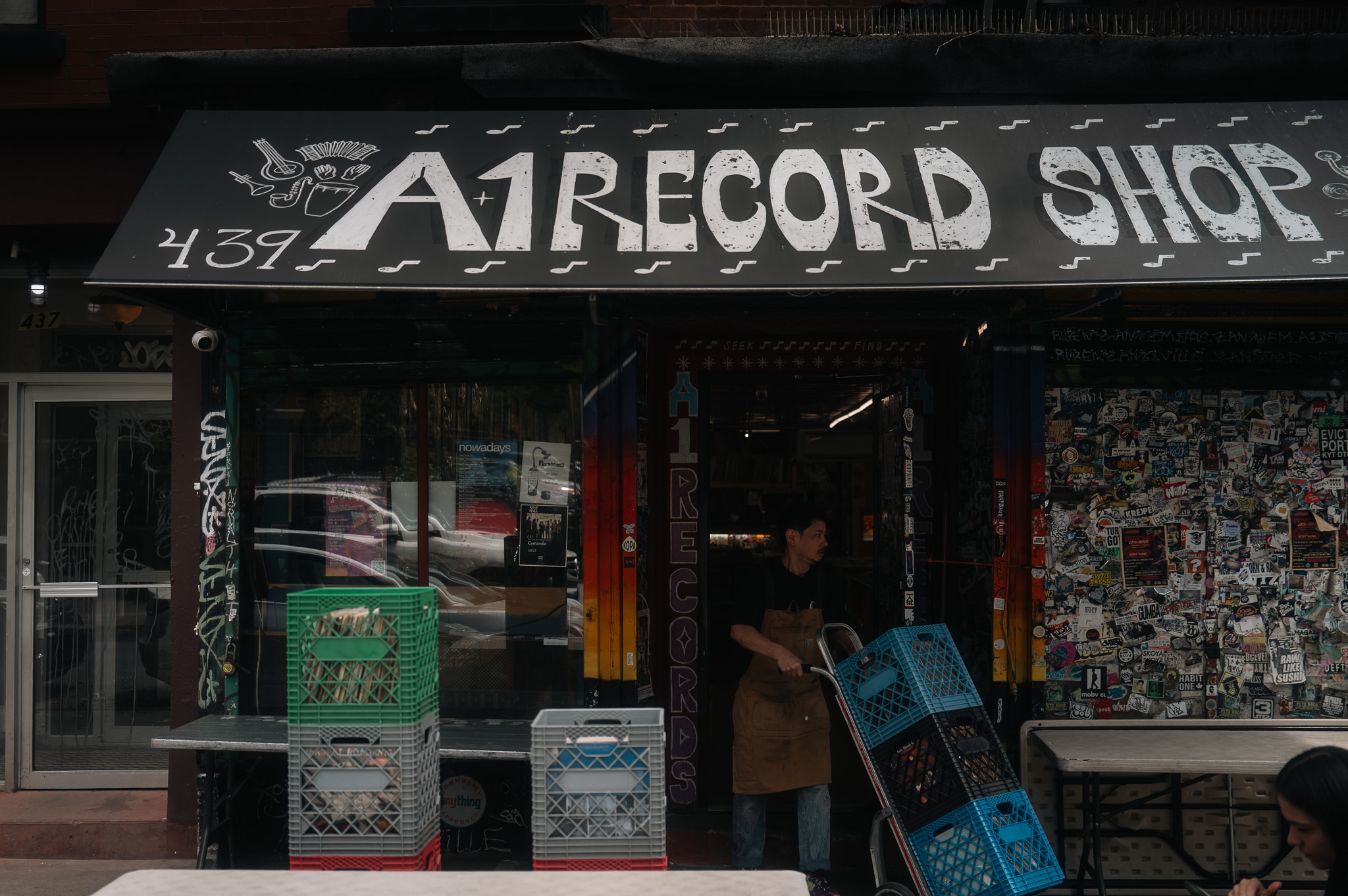 The A1 Record Shop storefront — staff in apron emerging through the door with the dolly, crates stacked on the sidewalk.